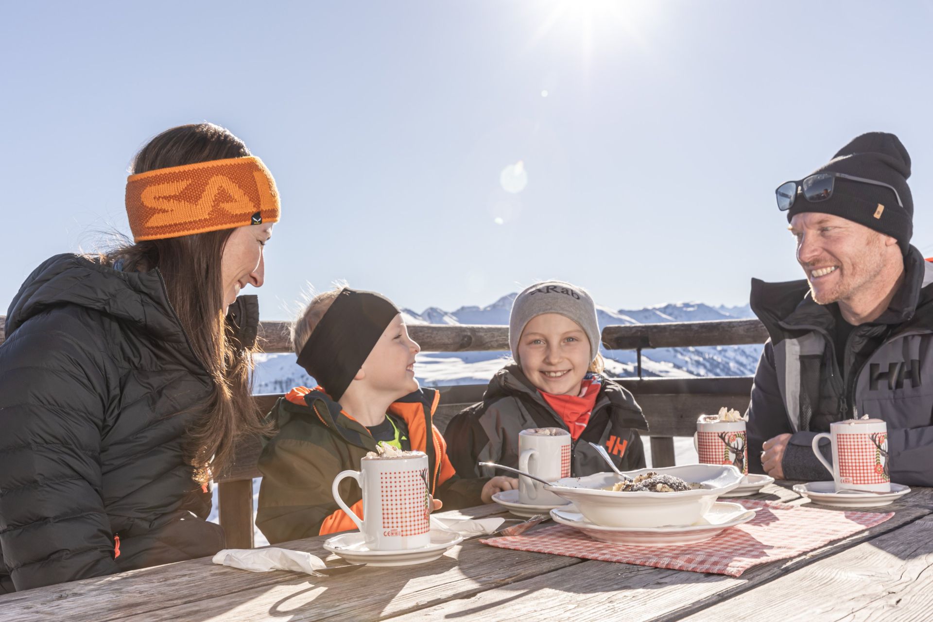 Familie mit heißer Schokolade auf einer sonnigen Hüttenterrasse im Skigebiet